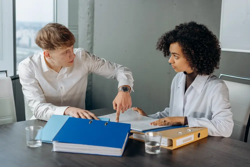 Two colleagues at a meeting table with binders; one points to a line in a printed document while the other follows along.