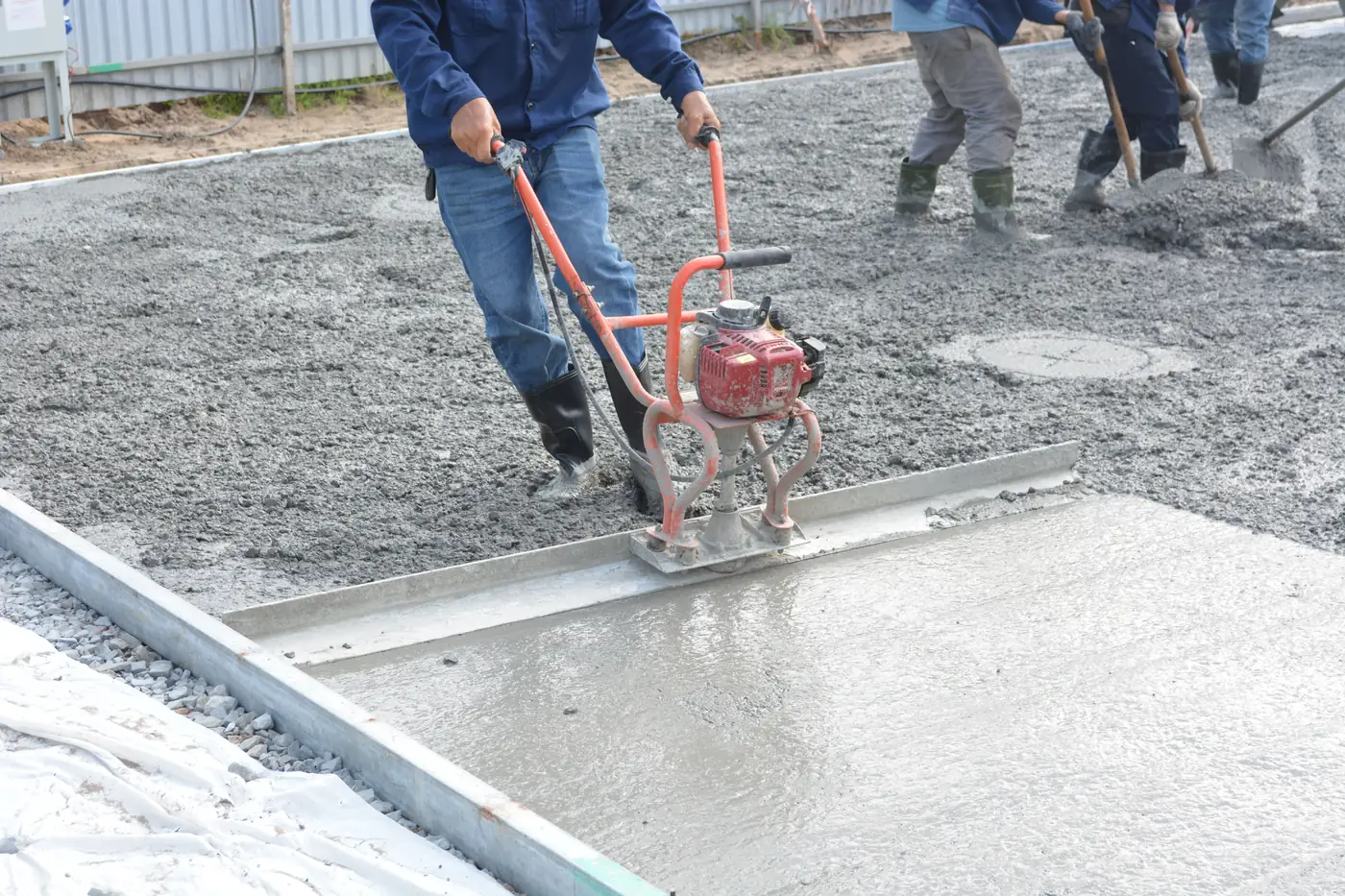 A concrete crew places and levels a fresh slab: one worker runs a power screed along the form while two others spread concrete behind him.