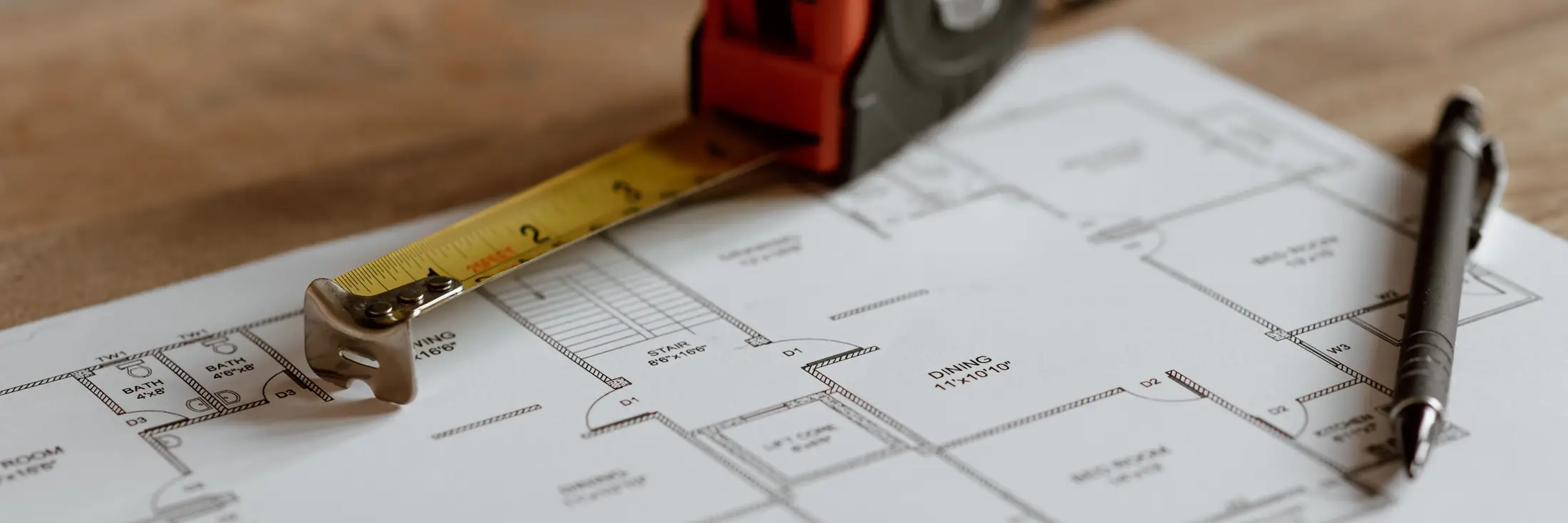A tape measure and a pen resting on a printed house floor plan on a wooden table.