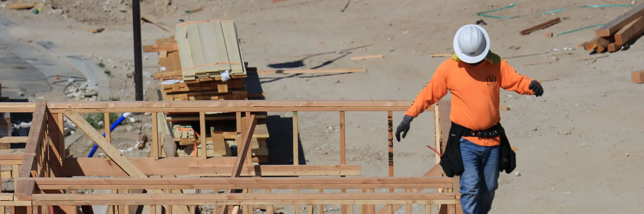 A framer in an orange shirt and white hard hat walks the top plate of new wall framing on a residential lot, with lumber stacked behind him.