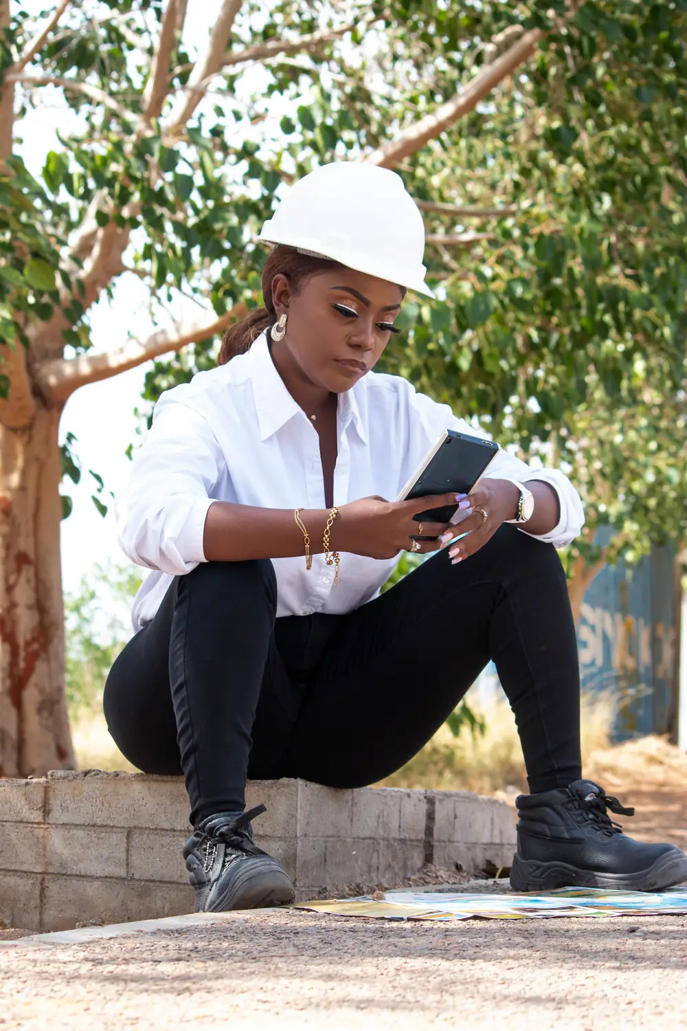 A woman in a white hard hat sits outdoors, reading her phone.