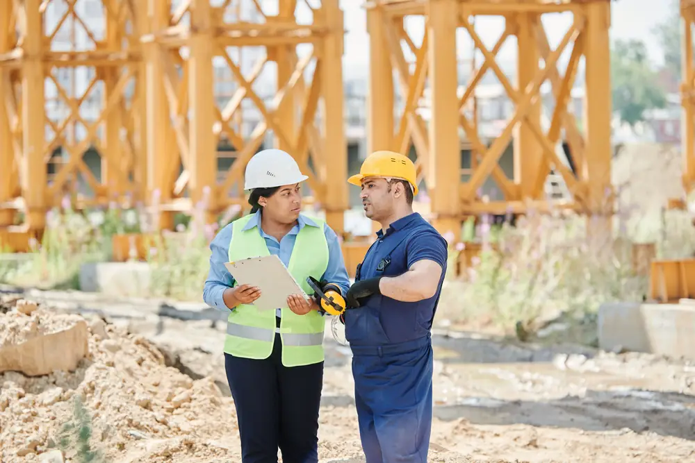 Two workers in hard hats talk on a jobsite; one holds a clipboard while the other points out an issue.