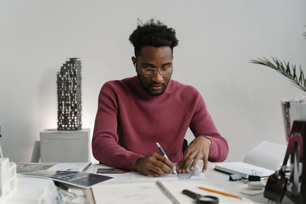 A man at a bright desk marks up a drawing with a pen, with plans, a notebook and a building model around him.
