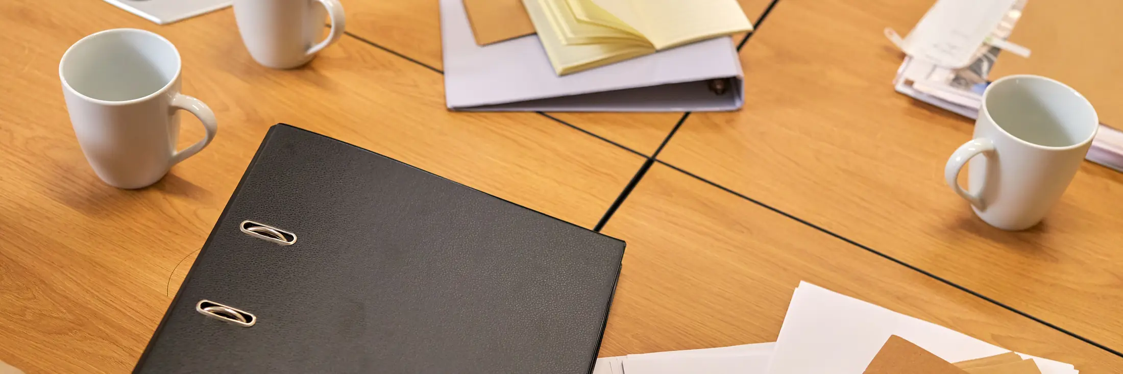 A meeting table after a working session: a black binder, notebooks, folders and coffee mugs.