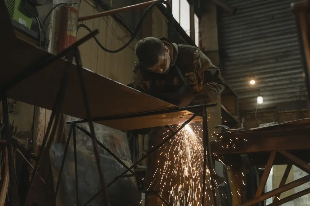 A metal worker bends over a steel sheet on a workbench as sparks fly from a grinder in a dim shop.