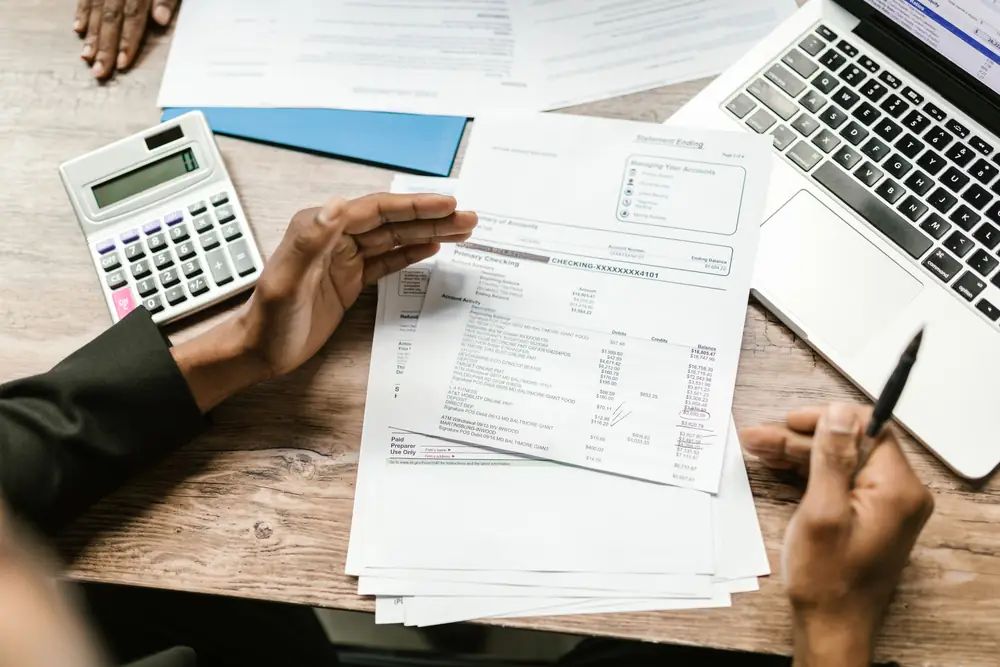 Hands going over a printed account statement at a wooden desk, with a calculator, a pen and an open laptop beside the paperwork.