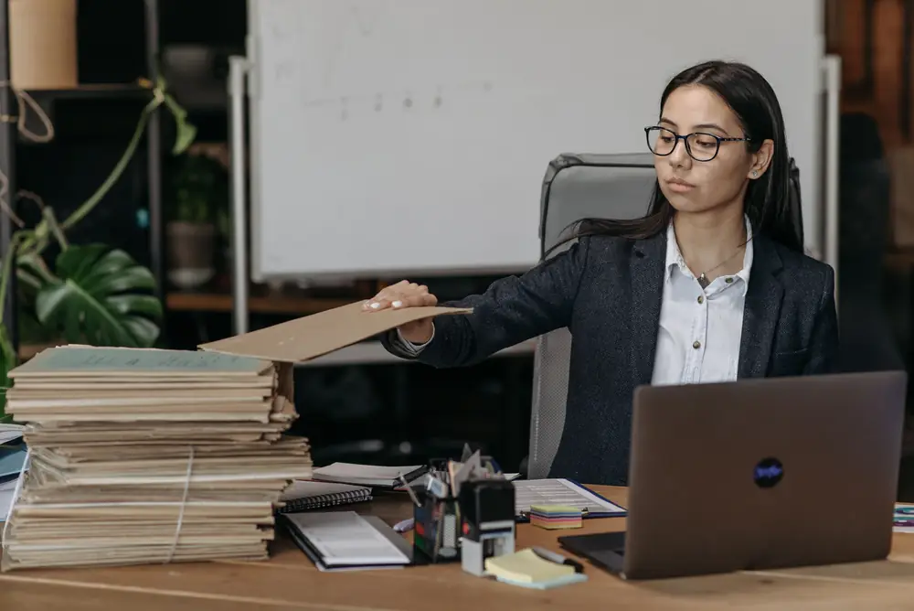 An office worker at a laptop reaches for a folder from a tall stack of paper files on her desk.
