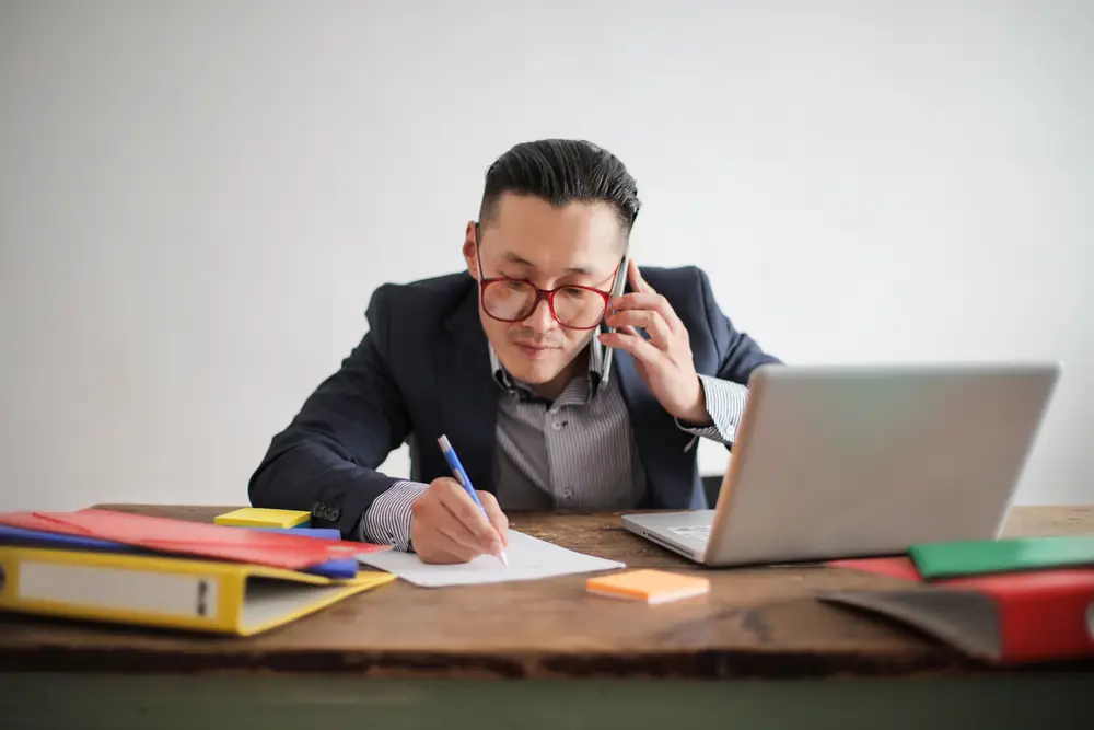 A man on a phone call takes notes at his desk, with a laptop and colored binders beside him.