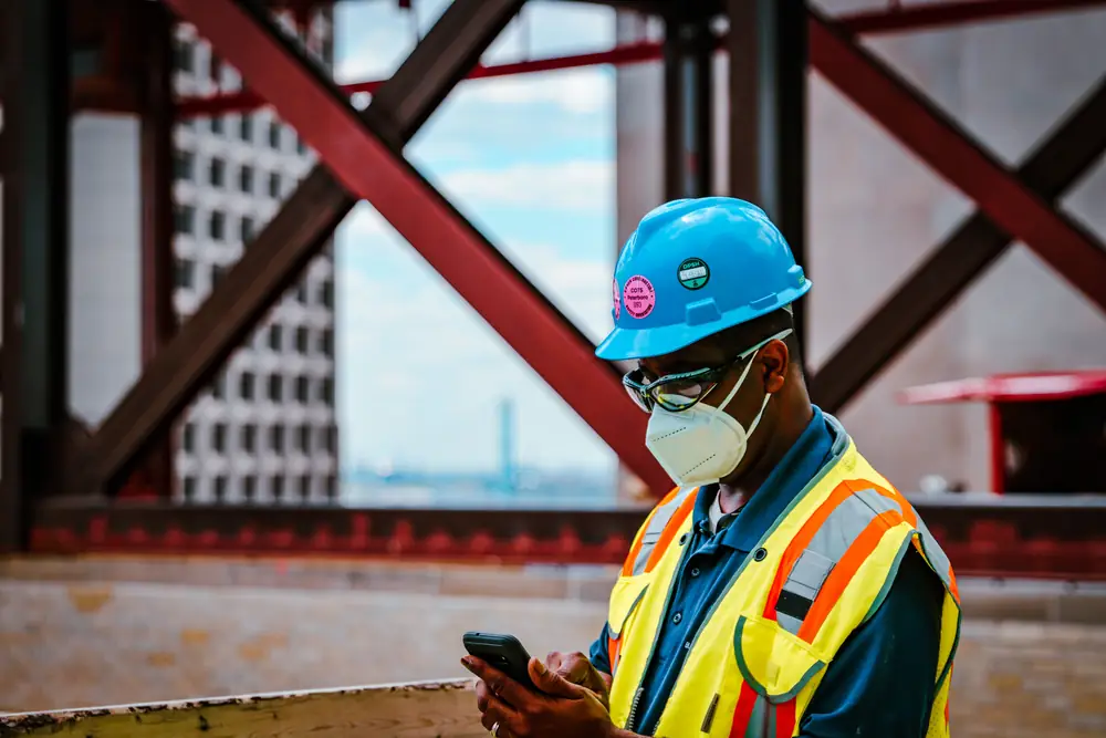 A site worker in a hard hat, safety glasses and a high-visibility vest checks his phone on a steel-frame jobsite.