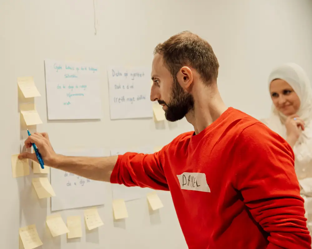 A man places sticky notes on a wall of handwritten sheets during a working session while a colleague watches.