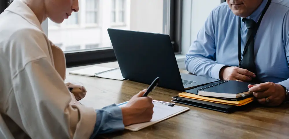 Two people at a table with a laptop; one takes notes on a clipboard while the other works from a stack of folders.