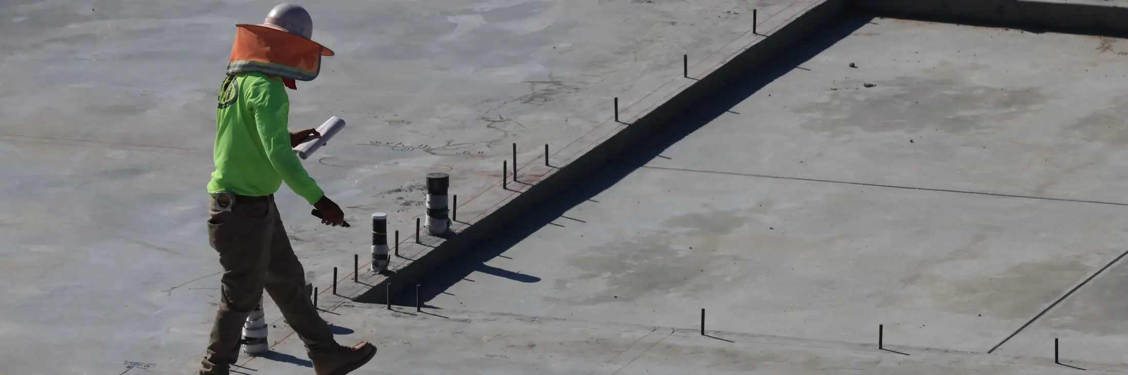 A worker walks across a fresh concrete slab in early light, past the anchor bolts along its edge.