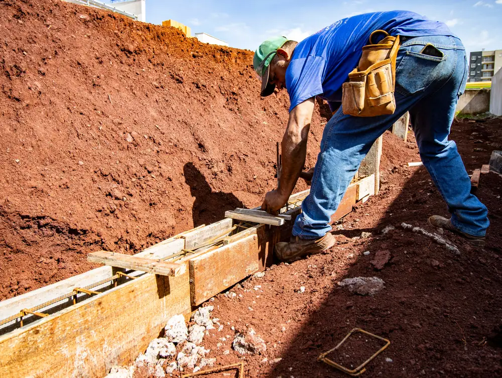 A worker in a tool belt sets wooden forms for a footing in a trench of red clay.